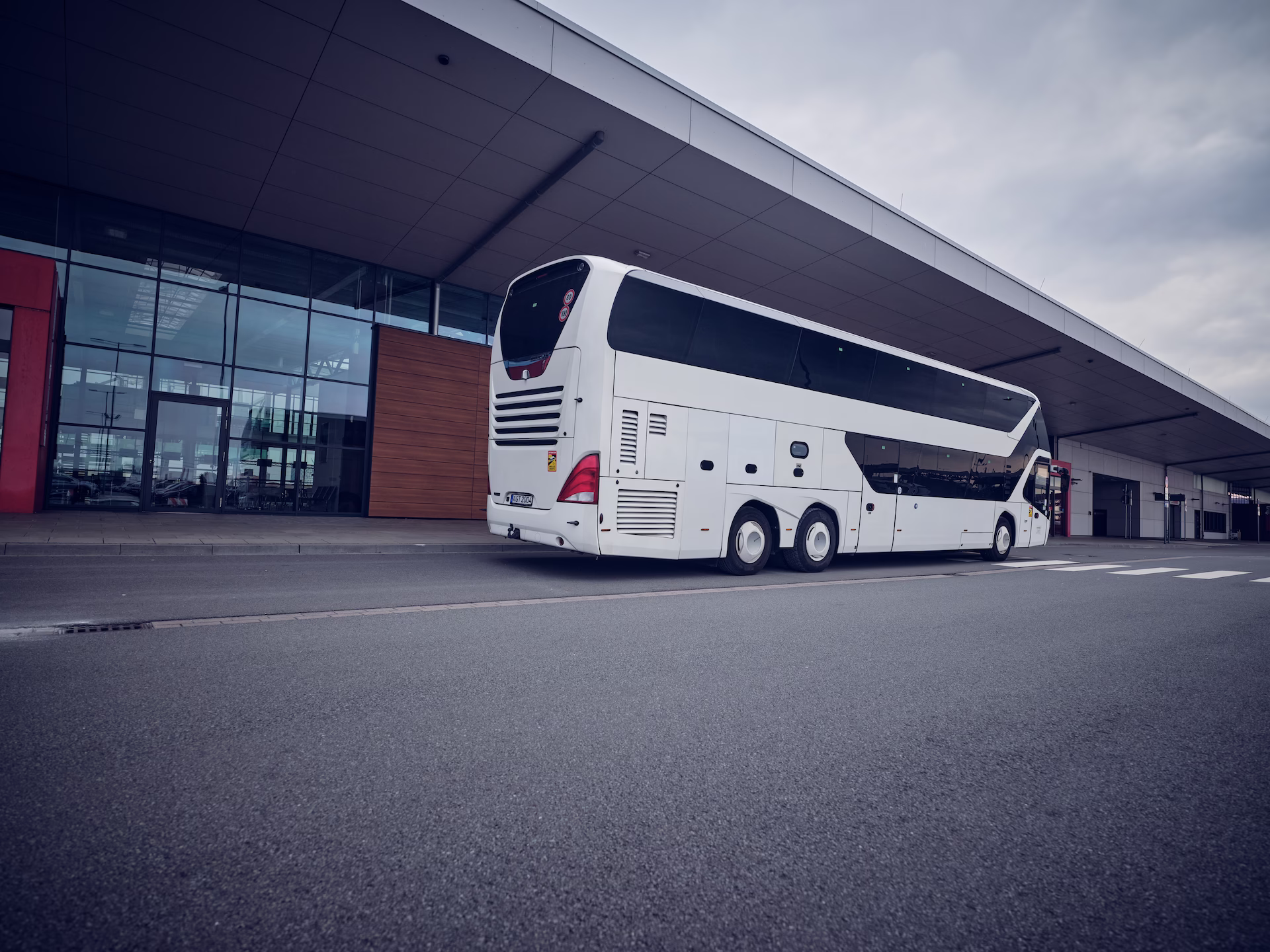 Ein großer, weißer Dreiachs-Doppelstockbus steht von schräg hinten fotografiert auf der Straße vor dem modernen Cruise Center Steinwerder in Hamburg unter dem weit auskragenden Dach des Terminals.