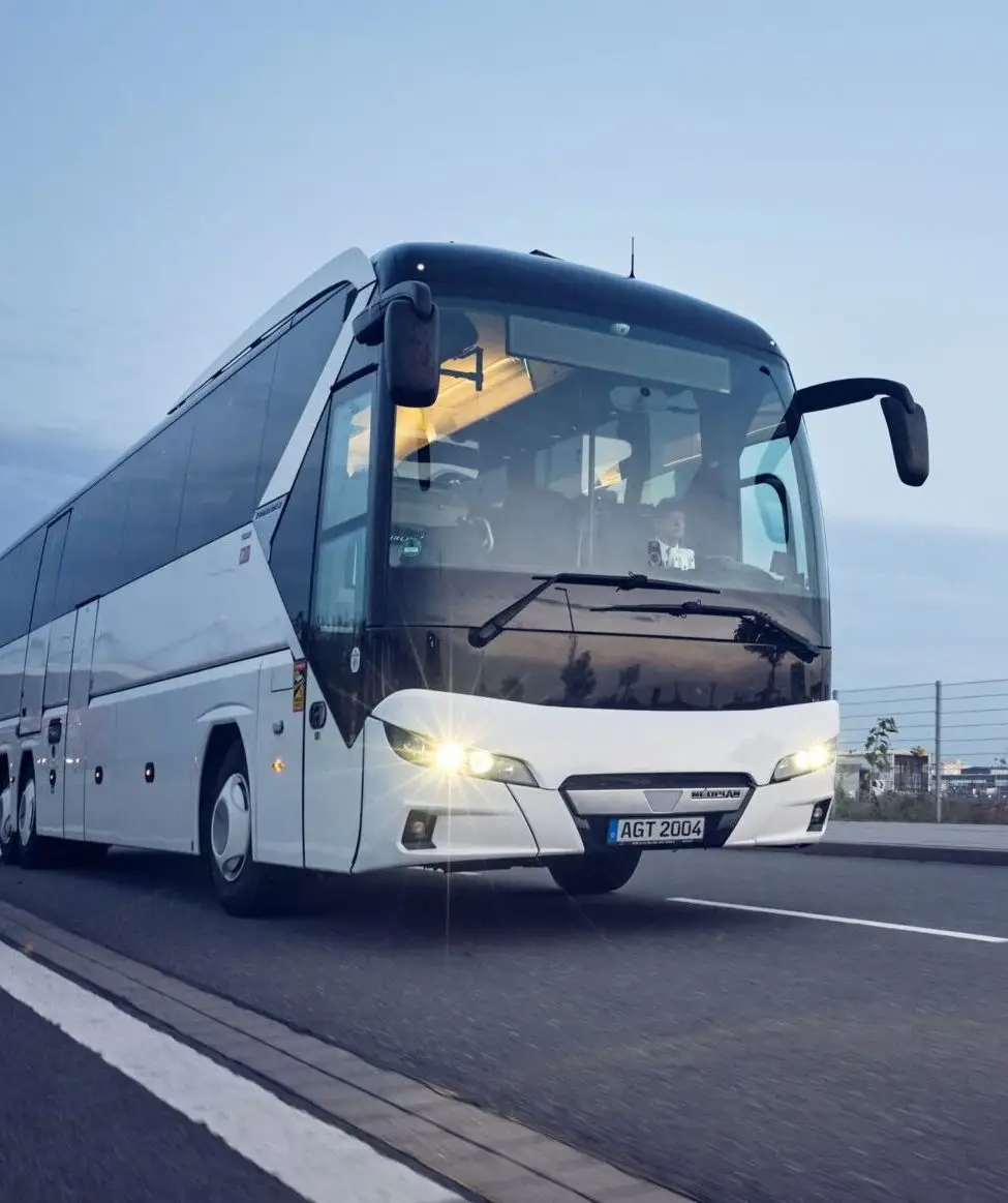 Ein weißer Neoplan-Reisebus mit eingeschalteten Scheinwerfern fährt bei Dämmerung auf einer asphaltierten Straße.