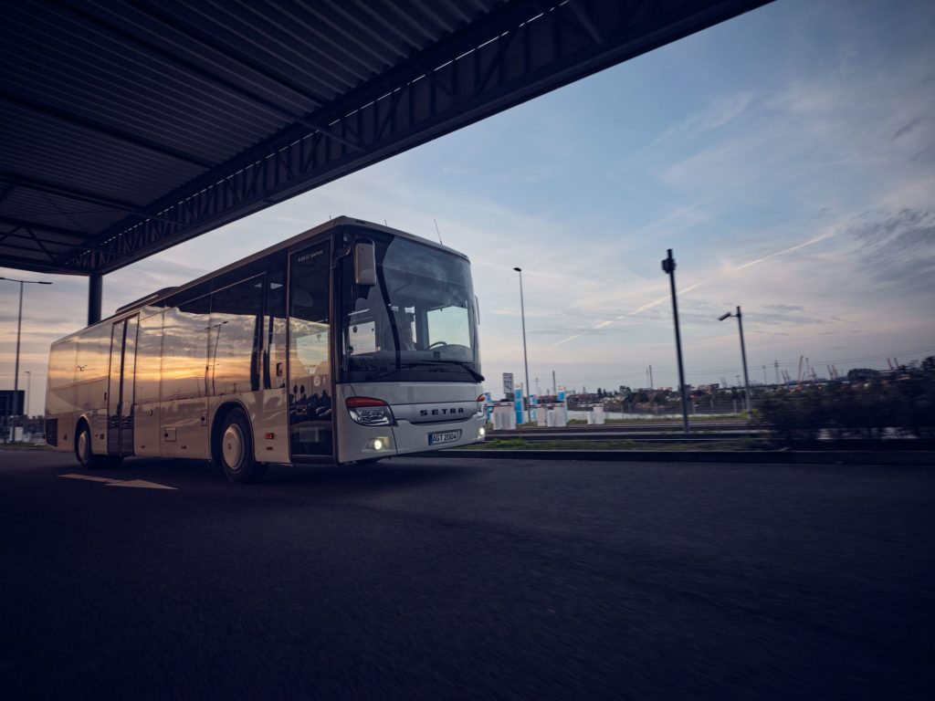 Weißer Linienbus fährt bei Sonnenuntergang unter einer Überdachung entlang.