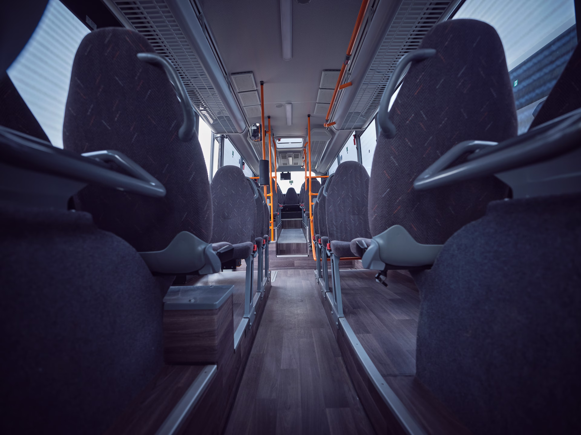 Blick von einer niedrigen Perspektive durch den Mittelgang eines Omnibusses nach vorne, flankiert von den Rückseiten grauer Polstersitze mit Haltegriffen und orangefarbenen Haltestangen im Hintergrund.
