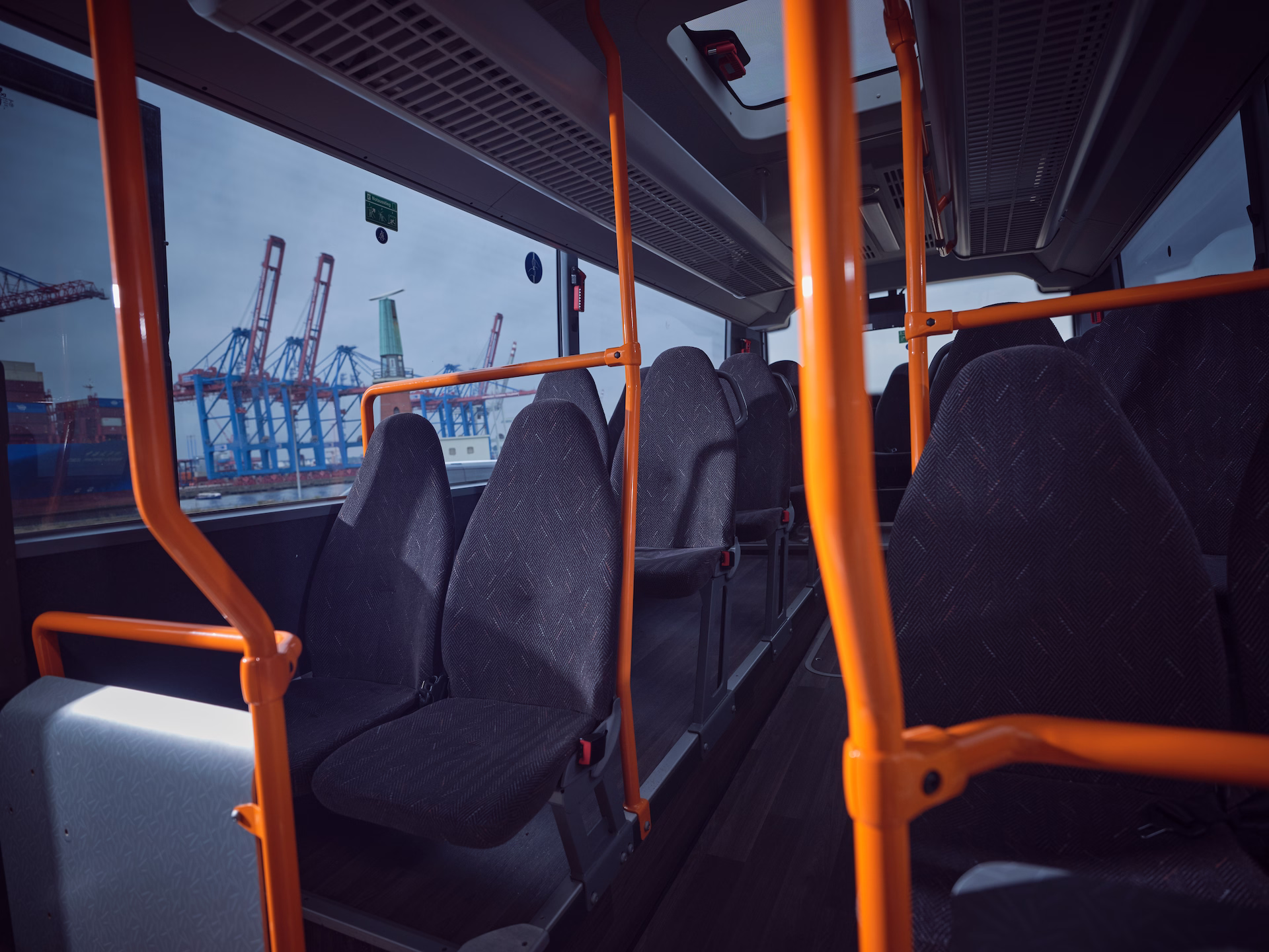Blick im Fahrgastraum eines Omnibusses auf graue Polstersitze und orangefarbene Haltestangen, mit deutlicher Sicht durch das linke Fenster auf die Containerkrane und die Lotsenstation im Hamburger Hafen.
