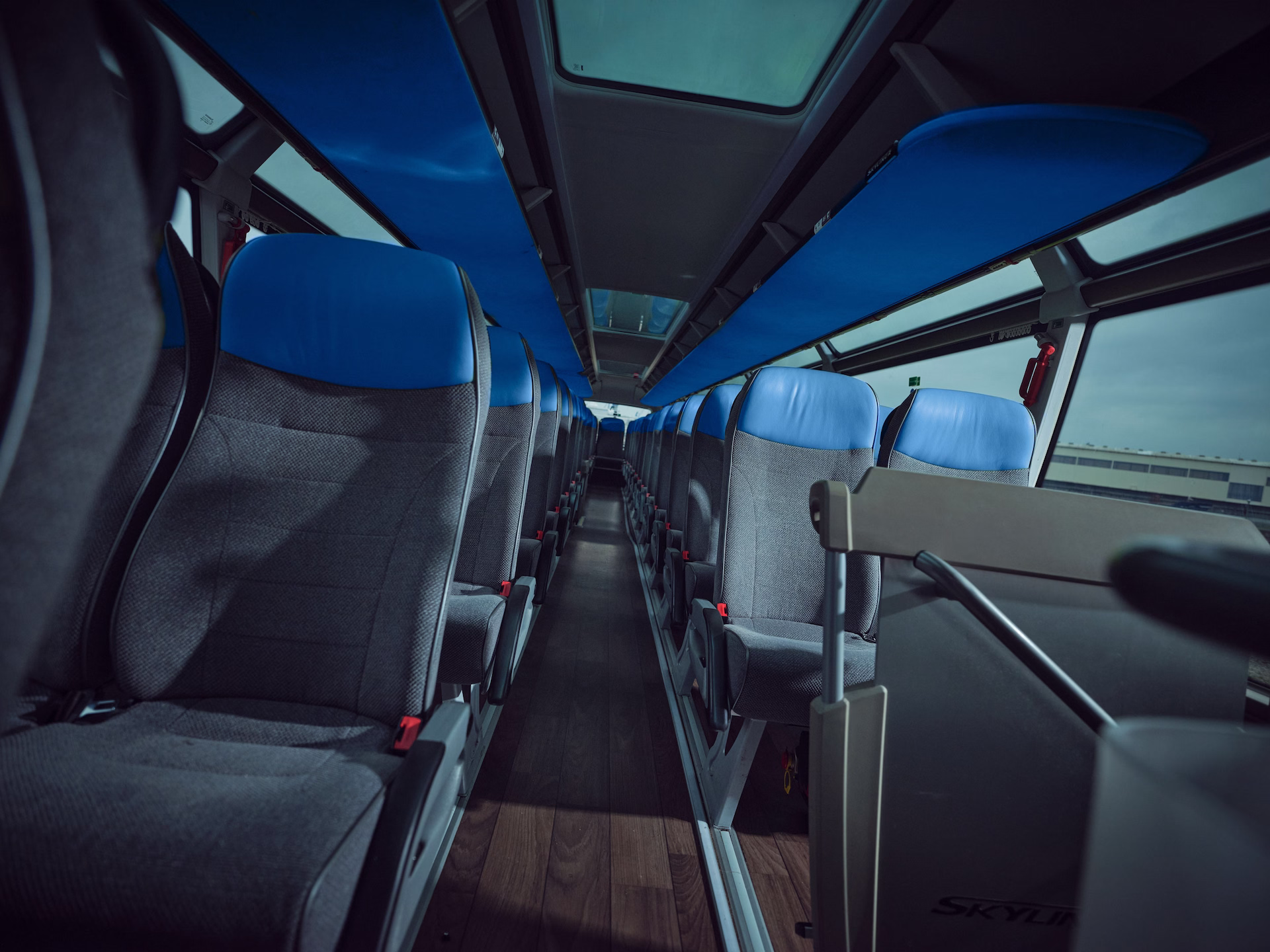 Blick durch den Mittelgang des Oberdecks eines Neoplan Skyliner Reisebusses mit grauen Sitzen und auffälligen blauen Kopfpolstern sowie einer blauen Verkleidung der Gepäckablagen unter der Decke.