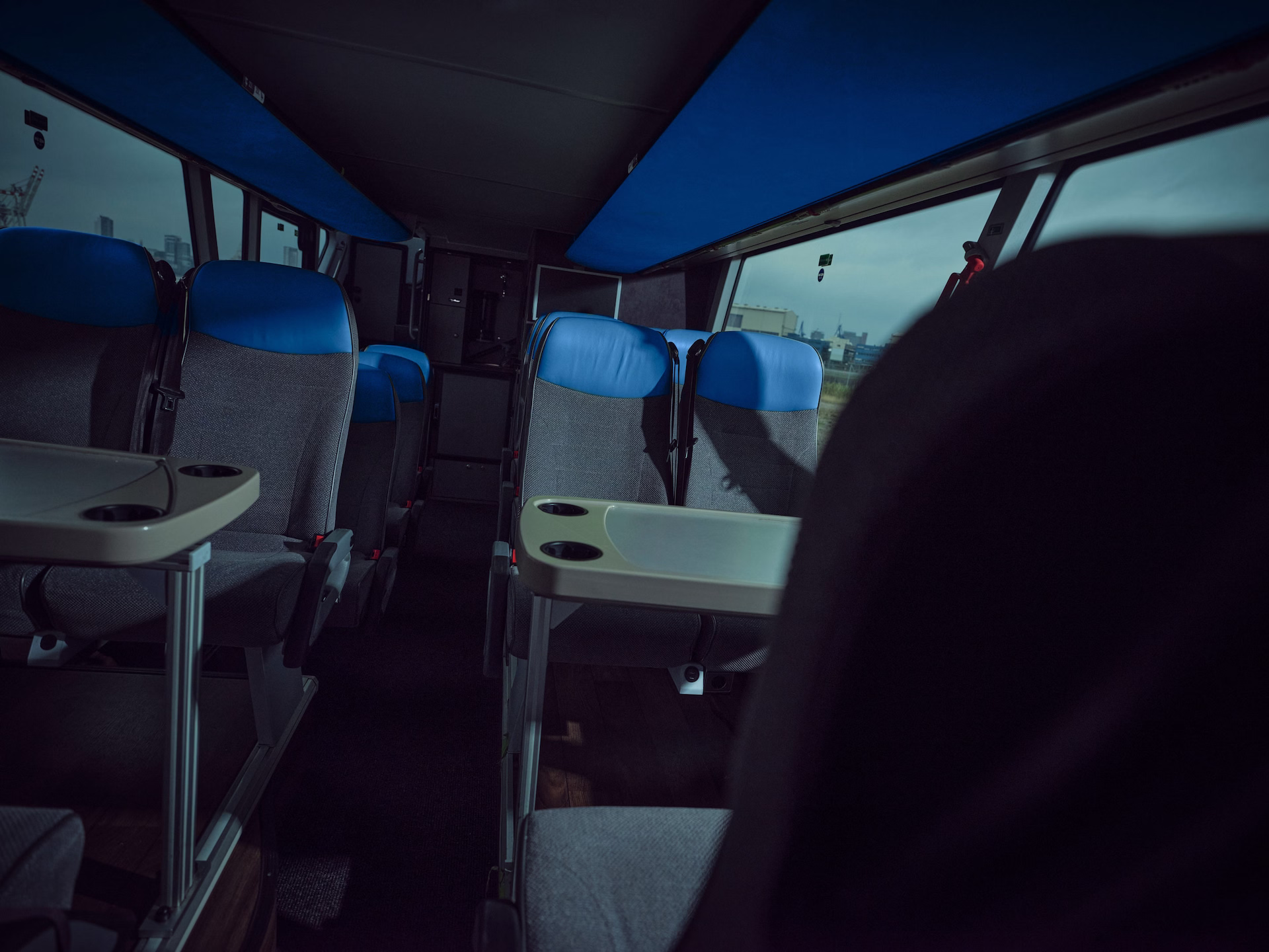 Blick in den Fahrgastraum eines Clubbusses oder Doppelstock-Unterdecks bei gedimmtem Licht, mit grauen Sitzen, die mit blauen Kopfpolstern versehen sind, und installierten Vis-à-vis-Tischen inklusive Becherhaltern.
