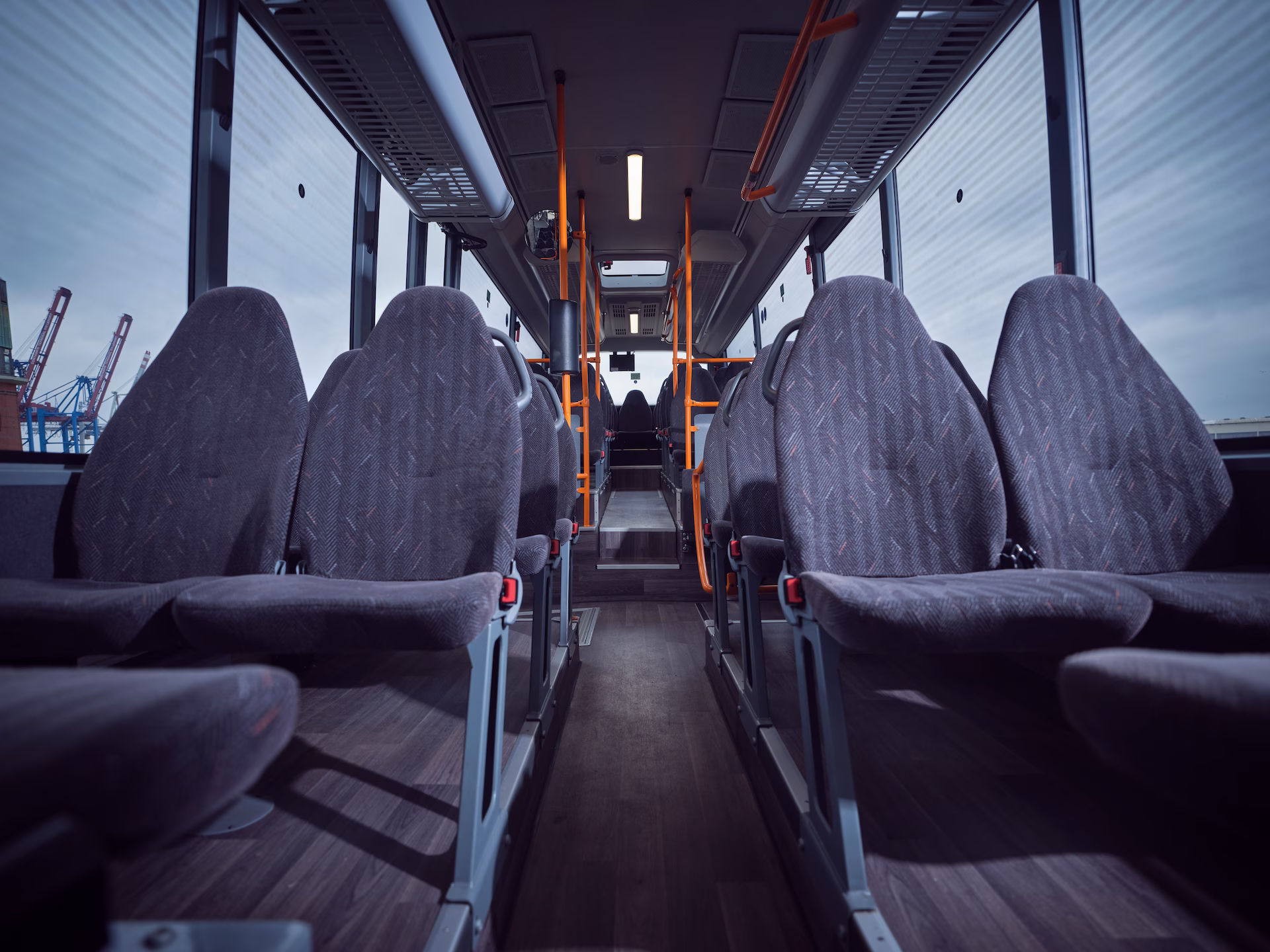 Symmetrischer Blick durch den Fahrgastraum eines Omnibusses mit Reihen aus grauen Polstersitzen und orangefarbenen Haltestangen, mit Blick auf Containerkrane im linken Hintergrund.