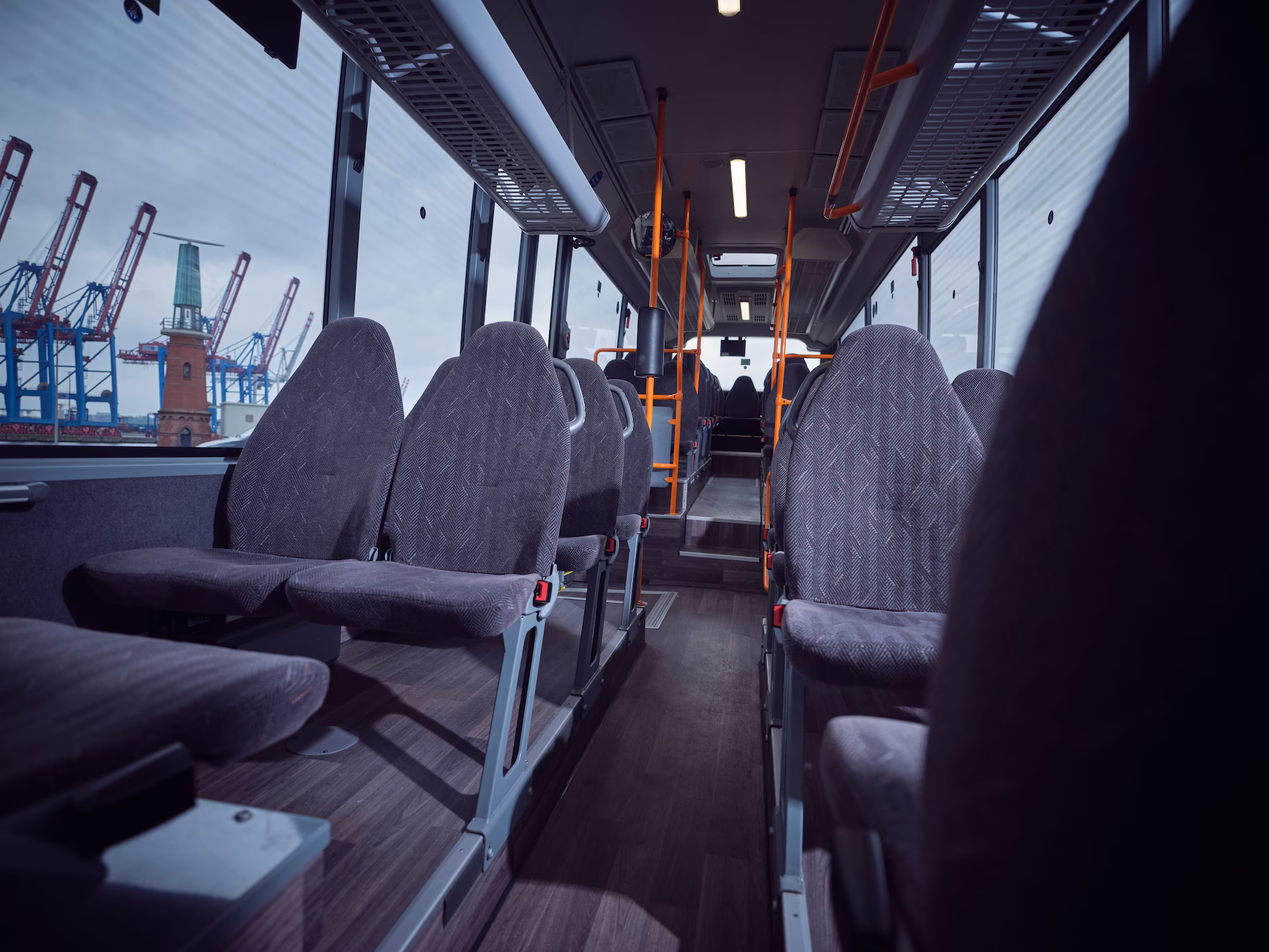 Blick durch den Fahrgastraum eines Omnibusses mit grauen Polstersitzen und orangefarbenen Haltestangen, mit Sicht durch das linke Fenster auf den Hamburger Hafen samt Containerkranen und Lotsenstation.
