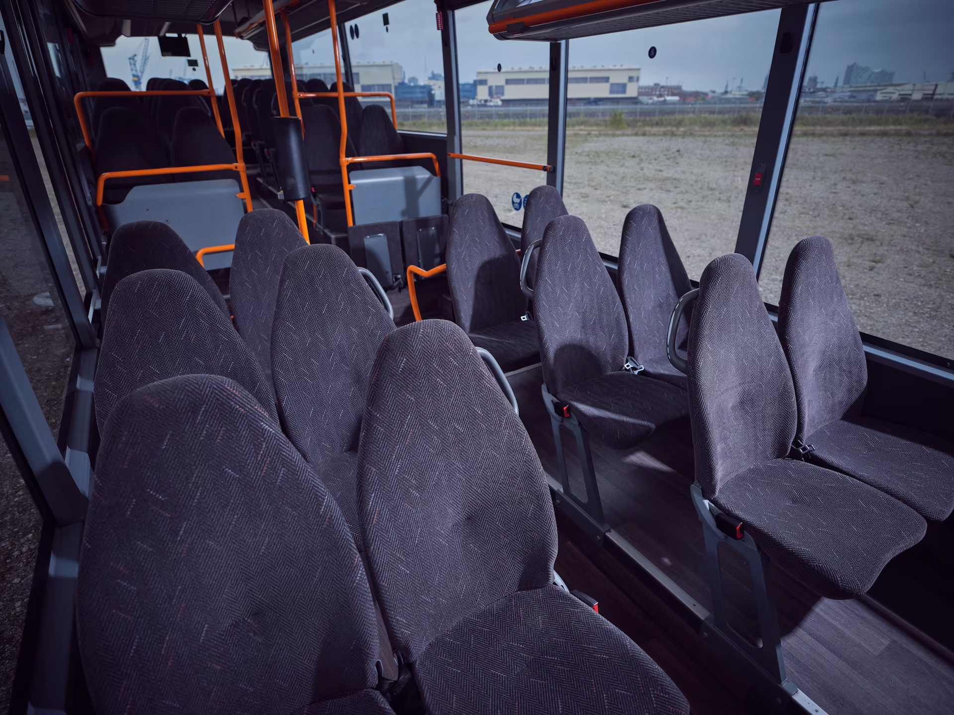 Blick in den Fahrgastraum eines Omnibusses mit Reihen aus dunklen, grauen Polstersitzen und orangefarbenen Haltestangen.