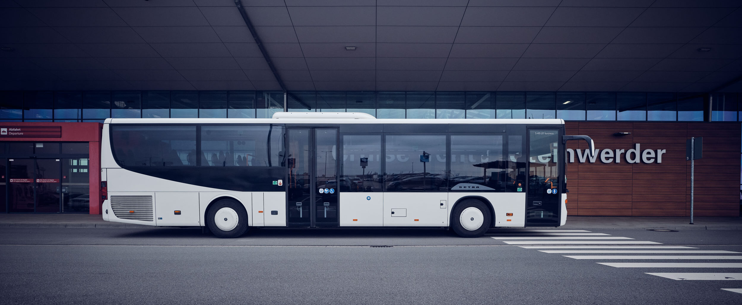 Ein weißer Shuttlebus steht vor einem modernen Terminalgebäude an einer ausgewiesenen Haltezone.