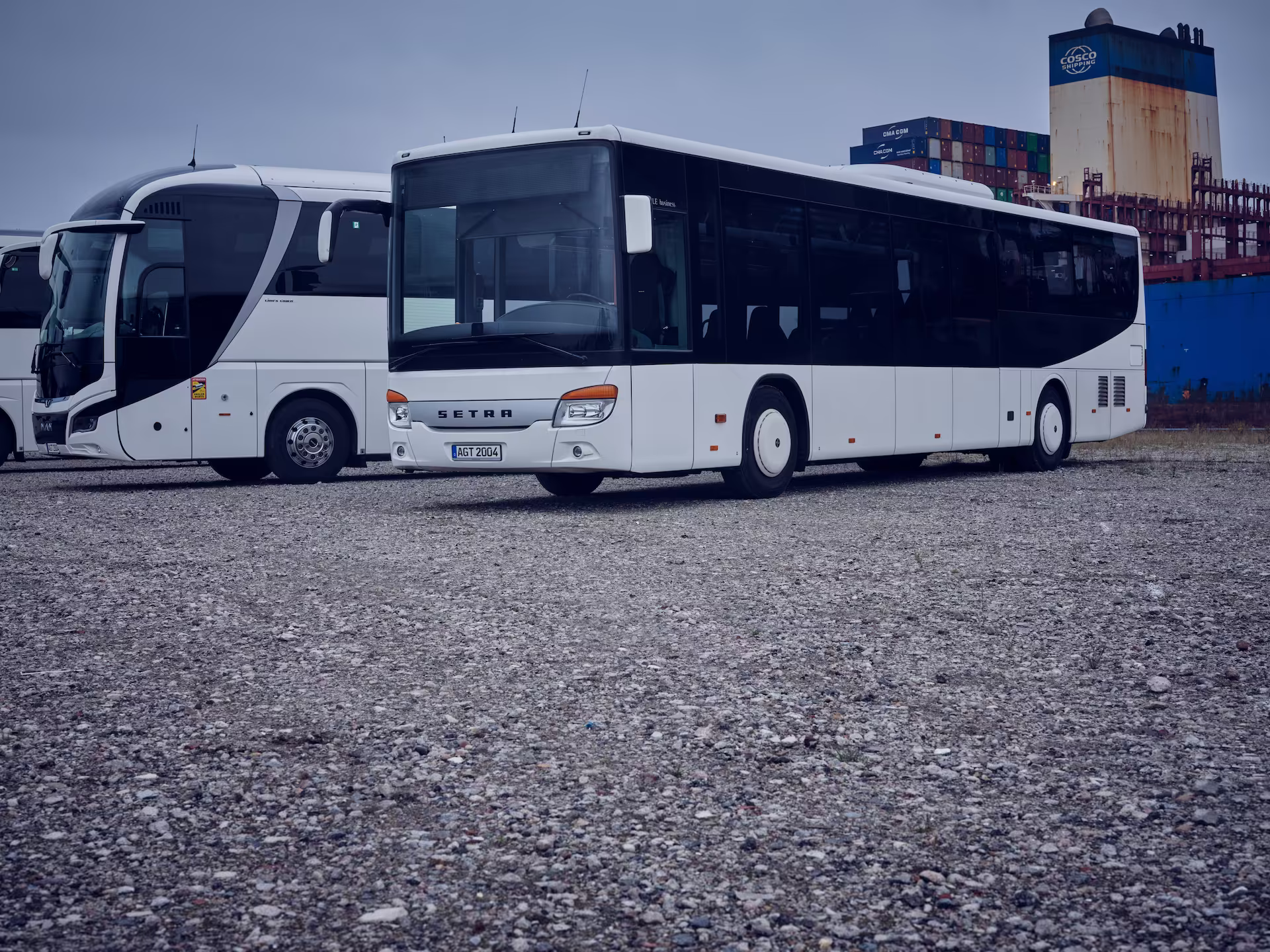Zwei weiße Busse aus der AGT-Fahrzeugflotte stehen auf einem Schotterplatz, mit Containern im Hintergrund unter einem bewölkten Himmel.