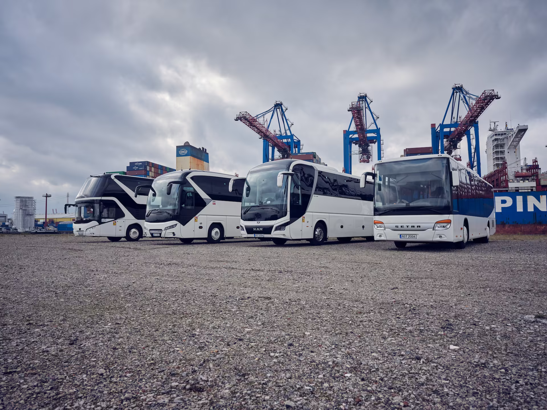 Vier Reisebusse stehen auf einer Freifläche vor einem Containerterminal.