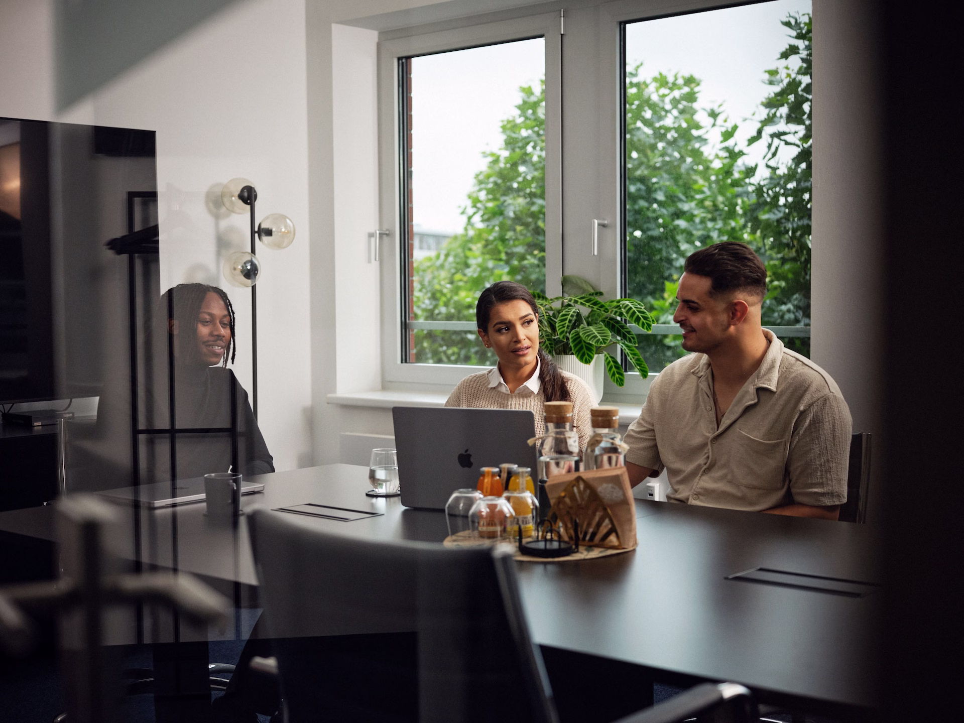 Drei Personen sitzen gemeinsam an einem Besprechungstisch und unterhalten sich vor einem Laptop in einem modernen, hellen Büro mit großen Fenstern und Pflanzen.