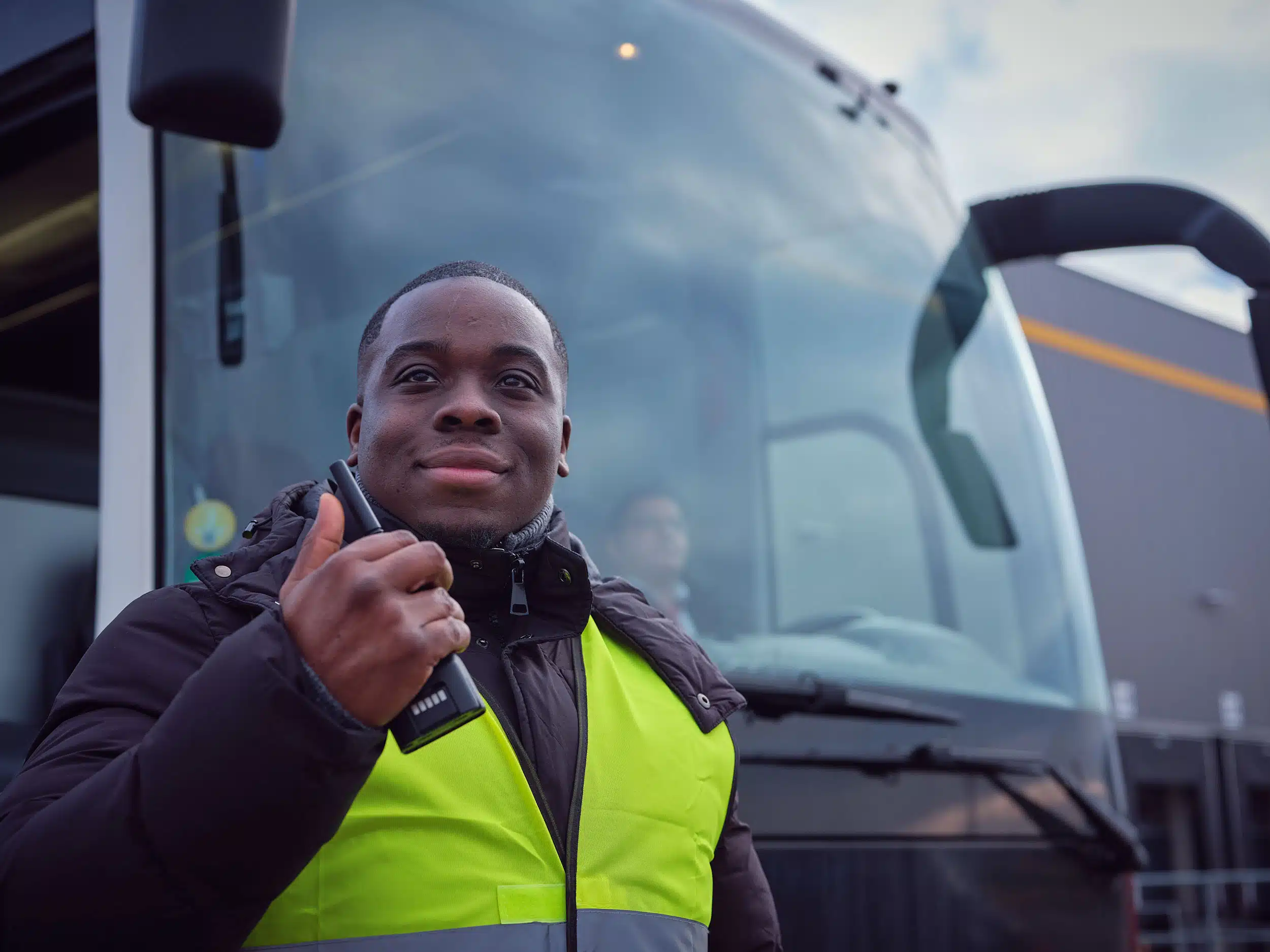 Ein Mann in einer neongelben Warnweste hält ein Funkgerät in der Hand und blickt lächelnd nach oben. Er steht direkt vor der großen Frontscheibe eines modernen Reisebusses.