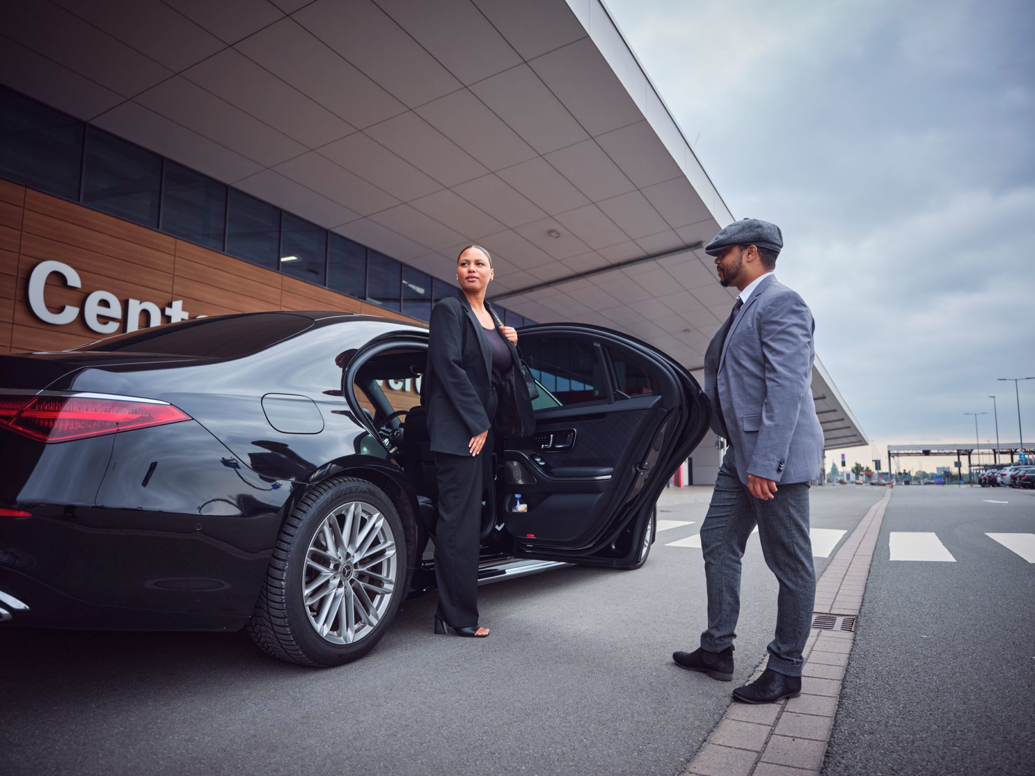 Eine Frau steigt vor einem Gebäude aus einer schwarzen Limousine aus, während ein Chauffeur neben der geöffneten Tür wartet.