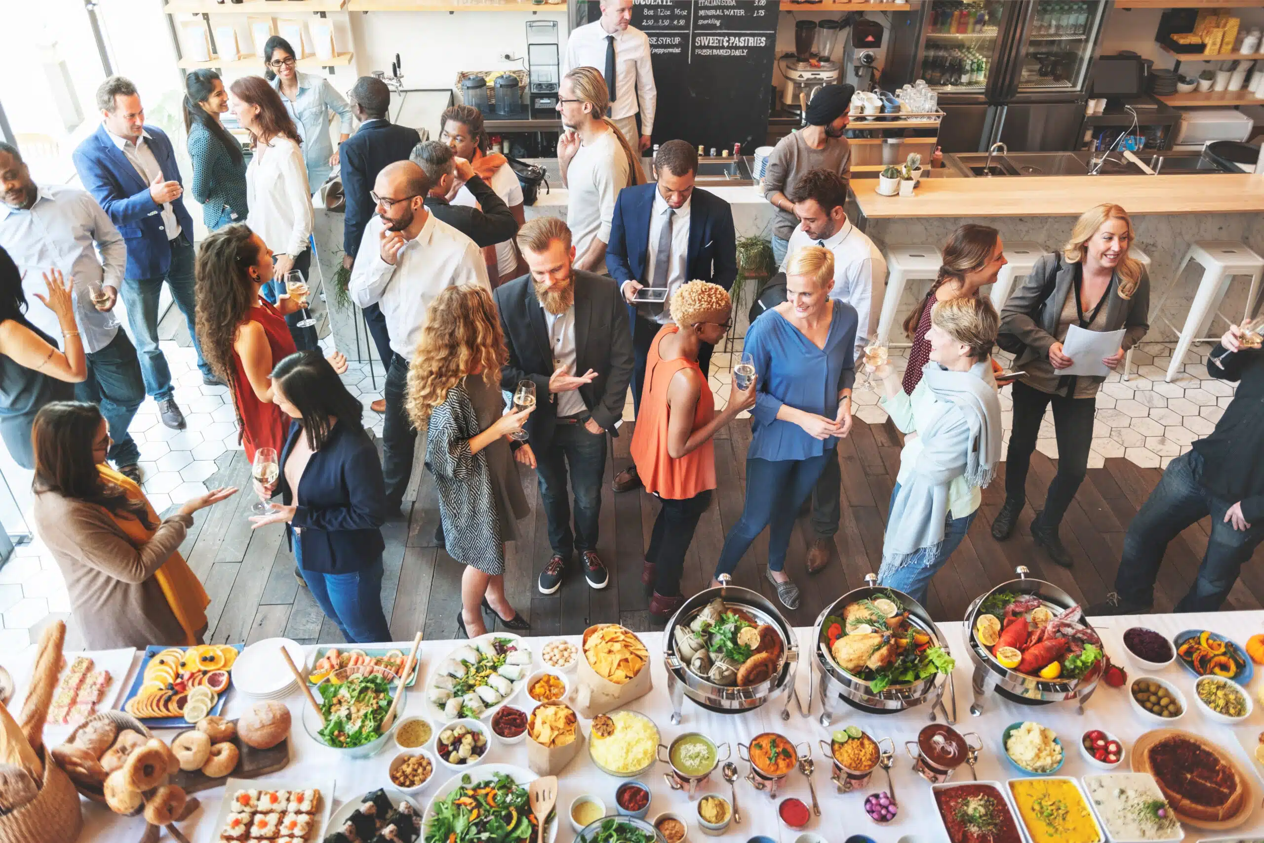 Vogelperspektive auf eine gut besuchte Netzwerk-Veranstaltung mit Gästen im Gespräch und einem langen Food-Buffet mit Salaten und warmen Speisen im Vordergrund.