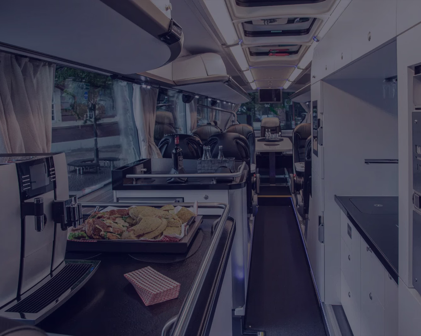 Blick in den Innenraum eines Reisebusses mit einer Kaffeemaschine und belegten Brötchen auf einer Theke im Vordergrund sowie einer weißen Küchenzeile und Sitzgruppen im hinteren Bereich.