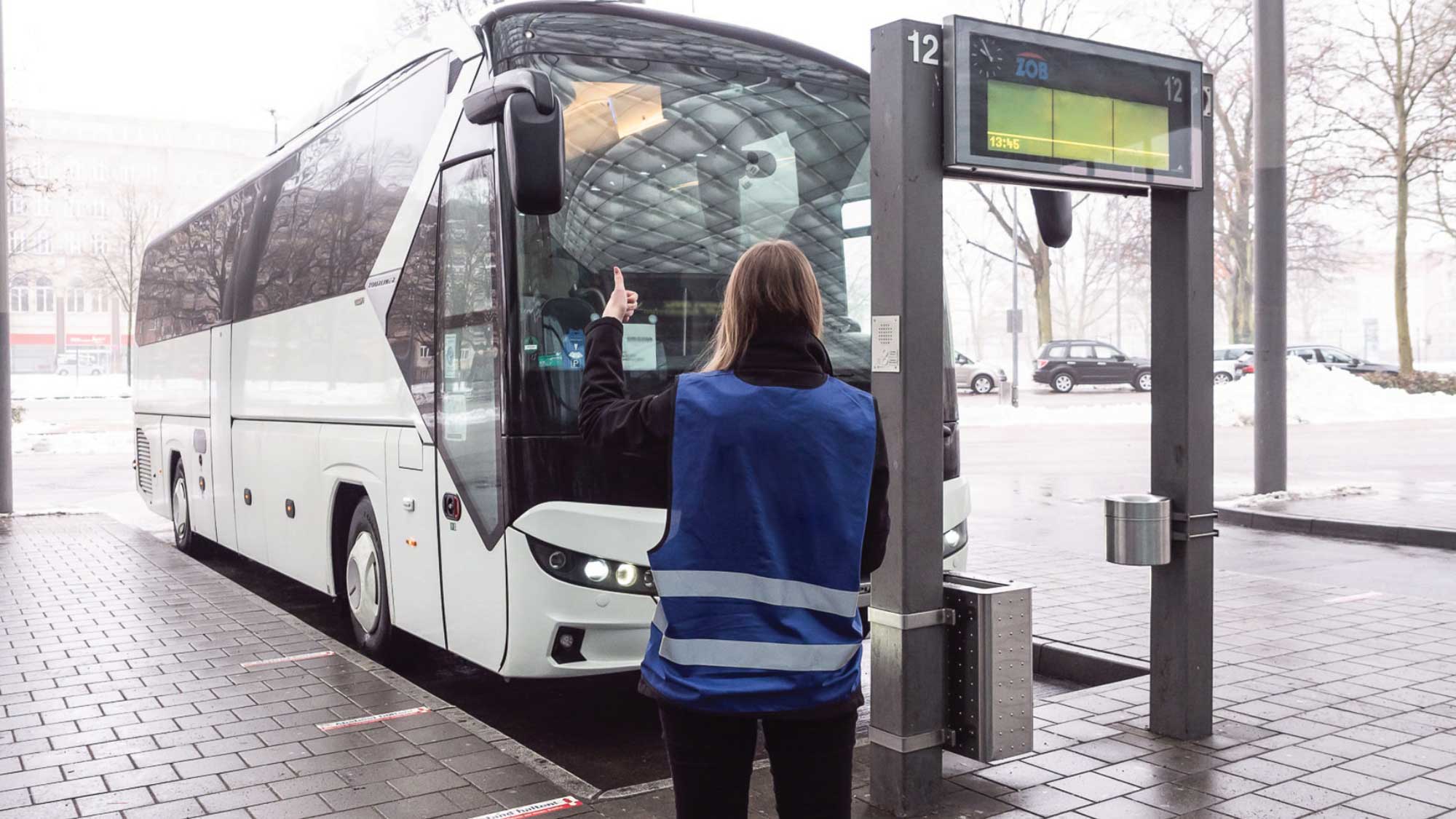 Eine Person in einer blauen Warnweste weist einen Reisebus an einem Bussteig ein. Der weiße Reisebus hält an einer Haltestelle mit digitaler Anzeige, während die Koordination der Ankunft vor Ort erfolgt.