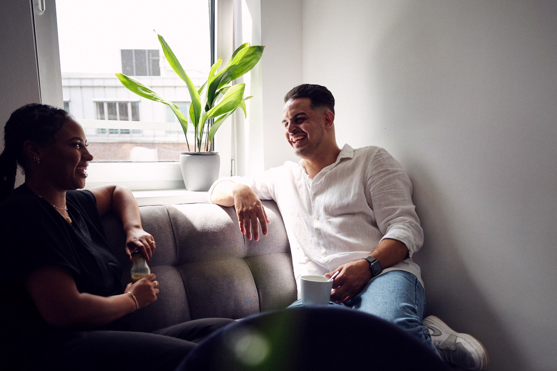 Zwei Personen sitzen auf einem Sofa nahe einem Fenster, unterhalten sich und lächeln. Auf der Fensterbank steht eine Topfpflanze.