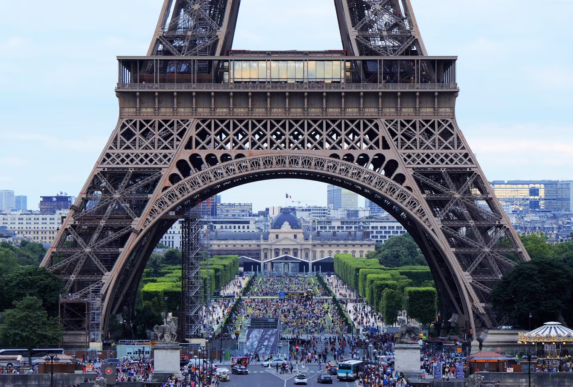 Blick durch den Eiffelturm auf die Parkanlage Champ de Mars in Paris mit zahlreichen Besucherinnen und Besuchern.