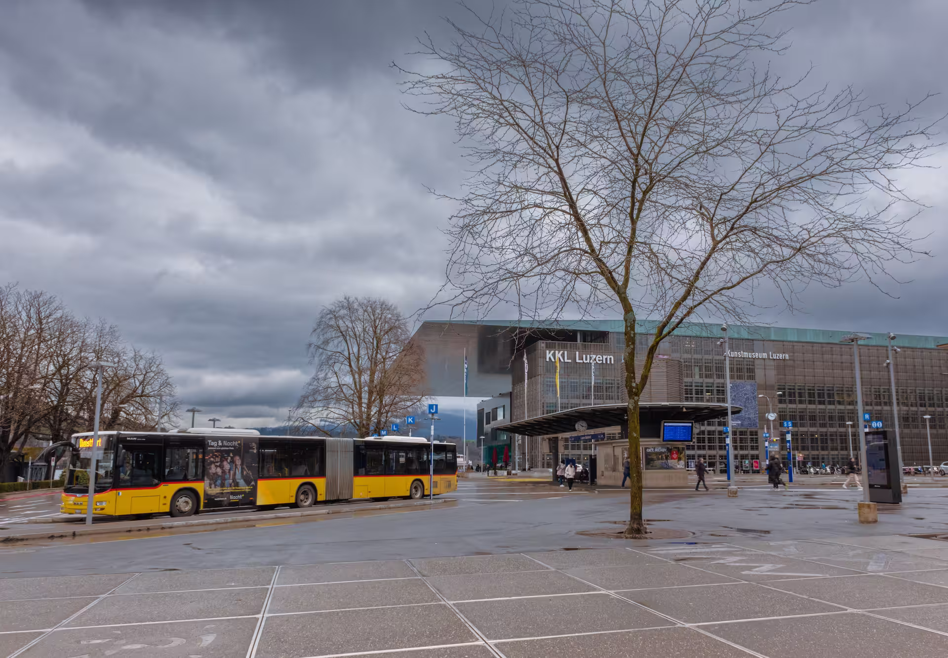 Gelber Gelenkbus an einer Haltestelle vor dem Kultur- und Kongresszentrum KKL Luzern bei bewölktem Himmel.