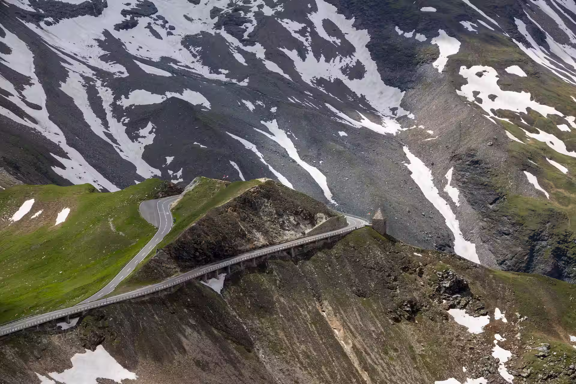 Kurvige Passstraße mit aufgeständertem Abschnitt an einem Berghang, umgeben von Geröllflanken und Schneefeldern im Hochgebirge.
