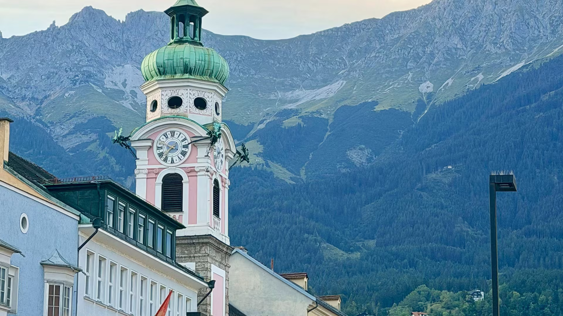 Turm der Spitalskirche in der Innsbrucker Innenstadt zwischen bunten Altbaufassaden, dahinter die Gipfel der Nordkette.