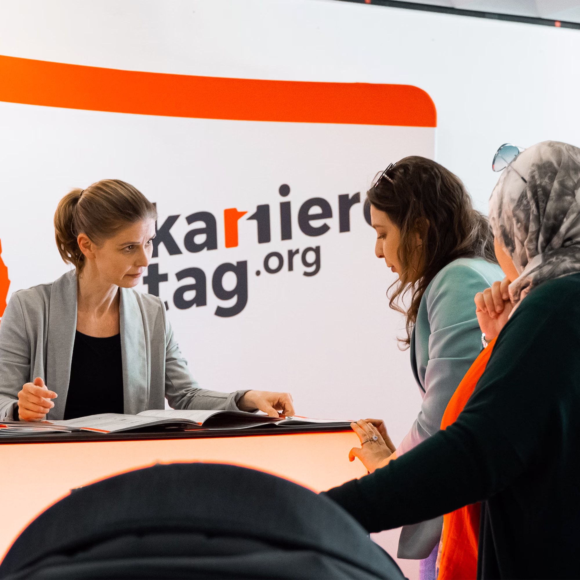 Mitarbeiterin berät zwei Besucherinnen an einem Messestand zum Thema Karriere und Ausbildung, vor einem Informationsbereich mit der Aufschrift „Karrieretag.org“.