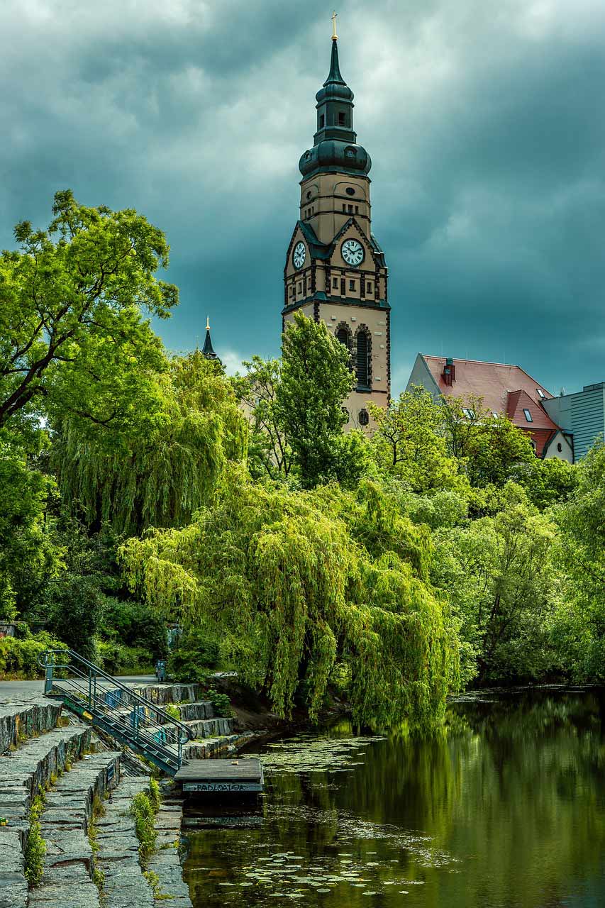 Kirchturm der Peterskirche in Leipzig erhebt sich über einer grünen Parklandschaft mit Teich und Weidenbäumen unter dramatisch bewölktem Himmel.