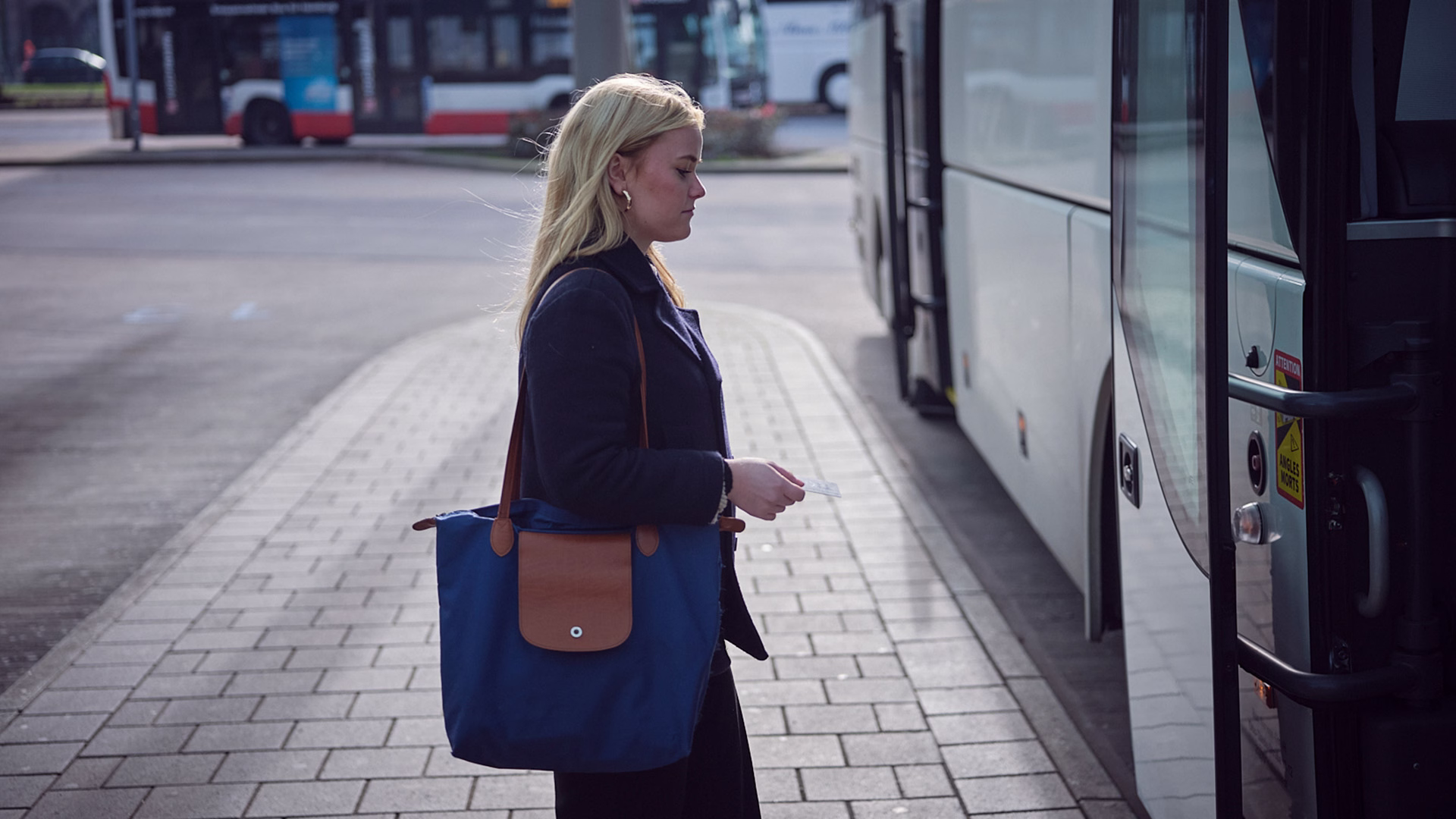Eine Frau mit blauer Tasche steigt an einer Haltestelle in einen Bus ein, während im Hintergrund ein weiterer Bus zu sehen ist.