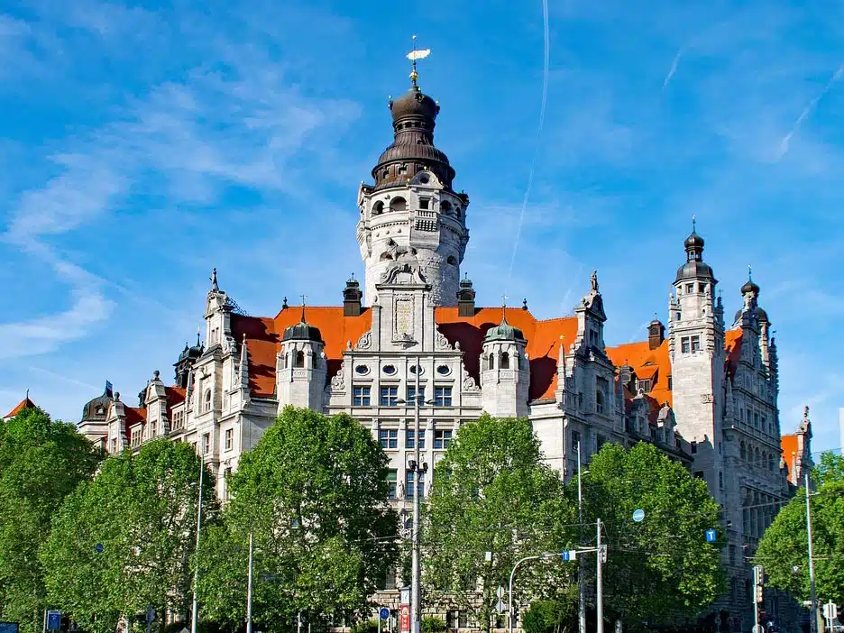 Historisches Neues Rathaus in Leipzig mit markantem Turm, rotem Dach und reich verzierter Fassade, umgeben von grünen Bäumen unter klarem blauem Himmel.
