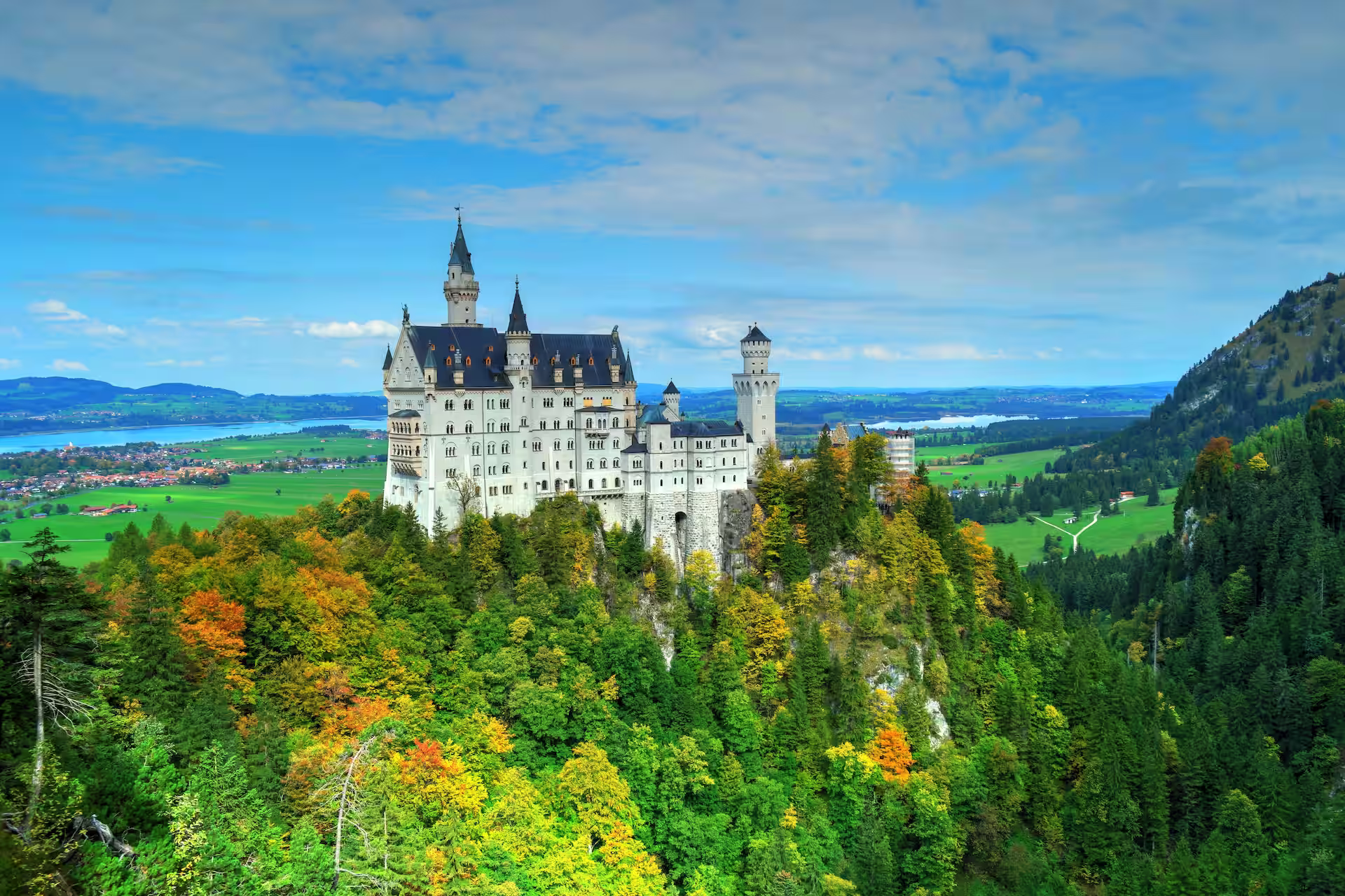 Schloss Neuschwanstein auf einem Felsen über herbstlich gefärbtem Wald, im Hintergrund das Allgäuer Voralpenland mit Seen.