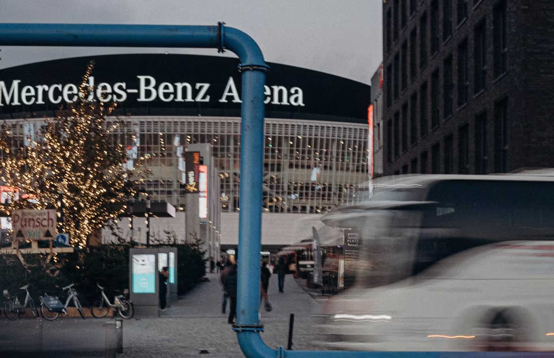 Reisebus fährt vor der Mercedes-Benz Arena in Berlin vorbei, während Besucherinnen und Besucher den beleuchteten Vorplatz in der Abenddämmerung nutzen.