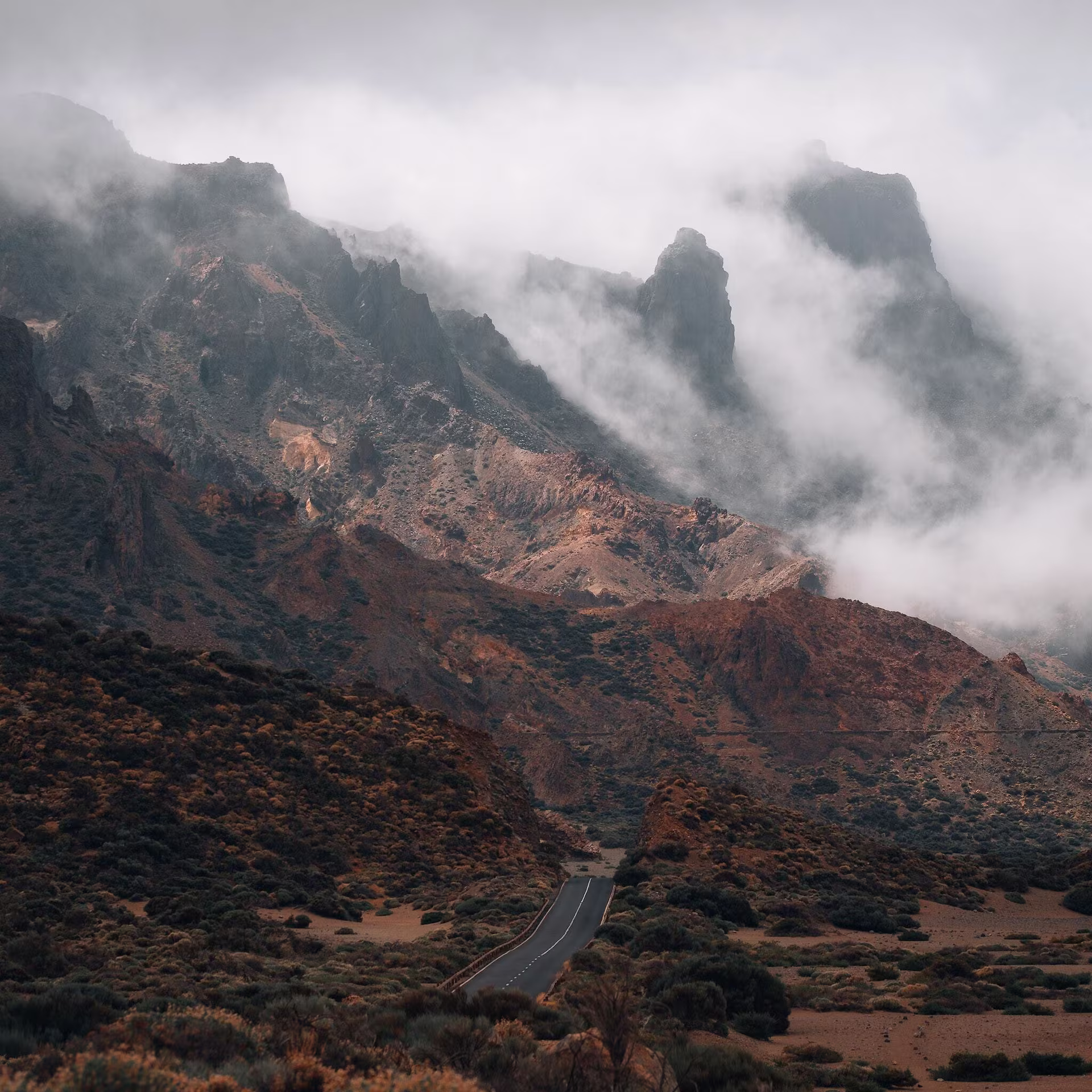 Schmale Straße führt durch eine felsige Berglandschaft, während Nebelschwaden und tiefe Wolken die Gipfel und Berghänge umhüllen.