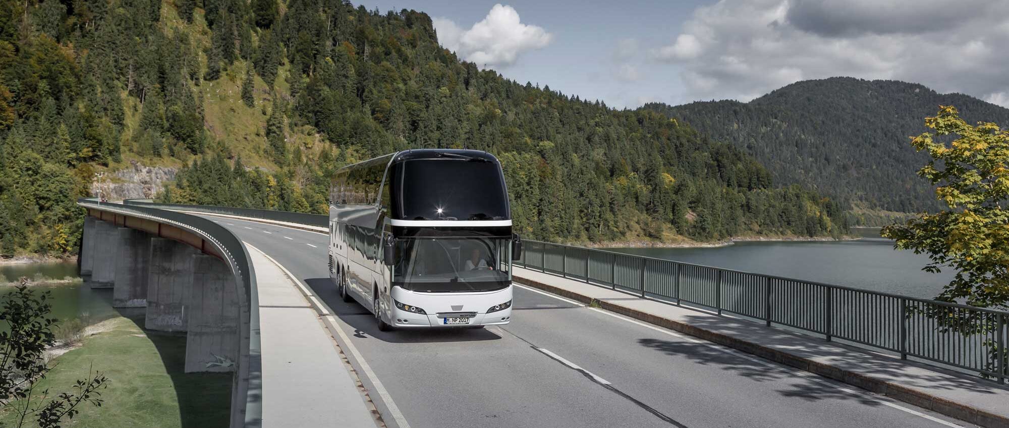 Reisebus auf einer Brücke entlang eines Sees in einer bergigen Waldlandschaft.