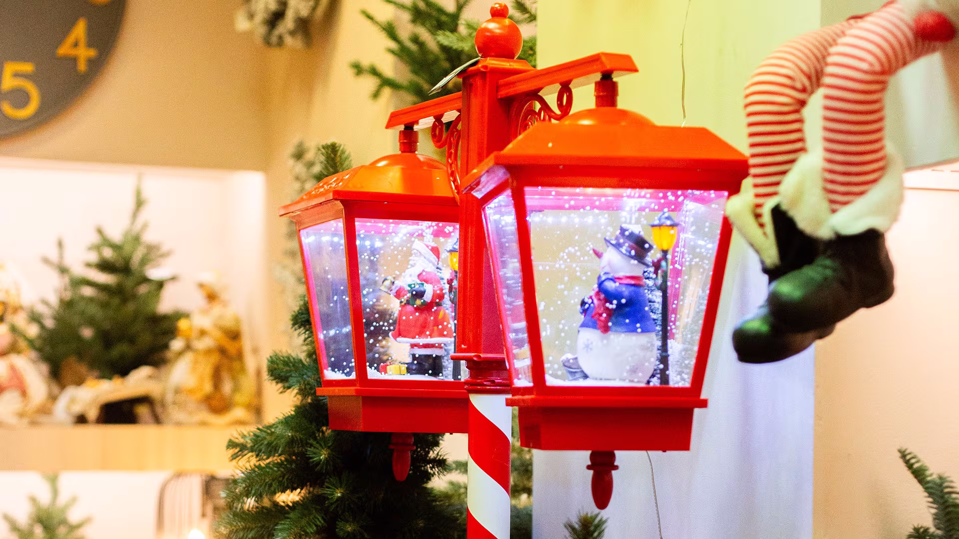 Rote Weihnachtslaterne mit Weihnachtsmann- und Schneemannfigur im künstlichen Schneegestöber, umgeben von Tannenzweigen und Weihnachtsdekoration.