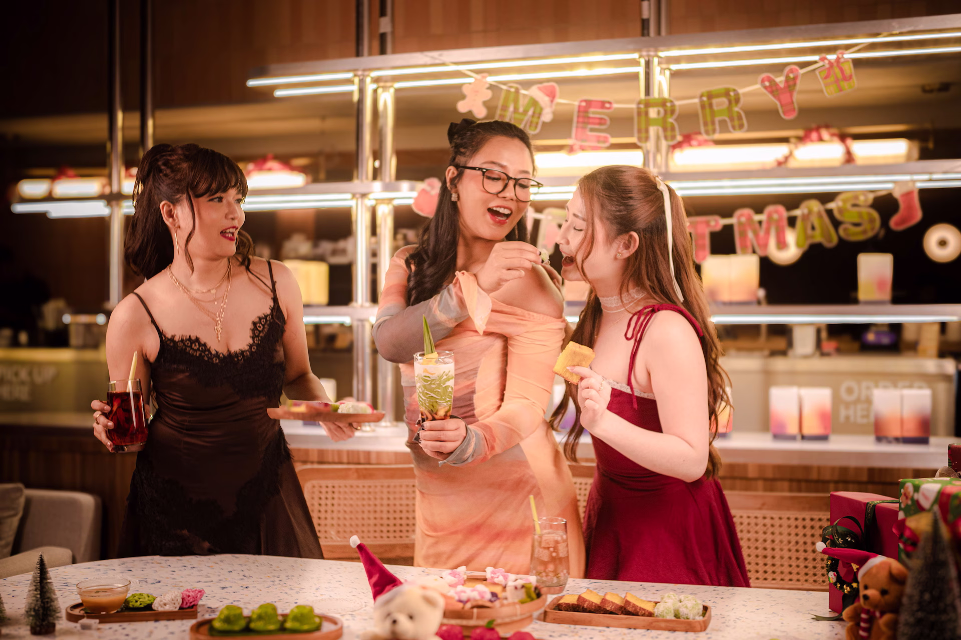 Drei Frauen in Abendkleidern feiern lachend an einer weihnachtlich dekorierten Bar mit Getränken, Gebäck und kleinen Snacks, im Hintergrund eine Girlande.