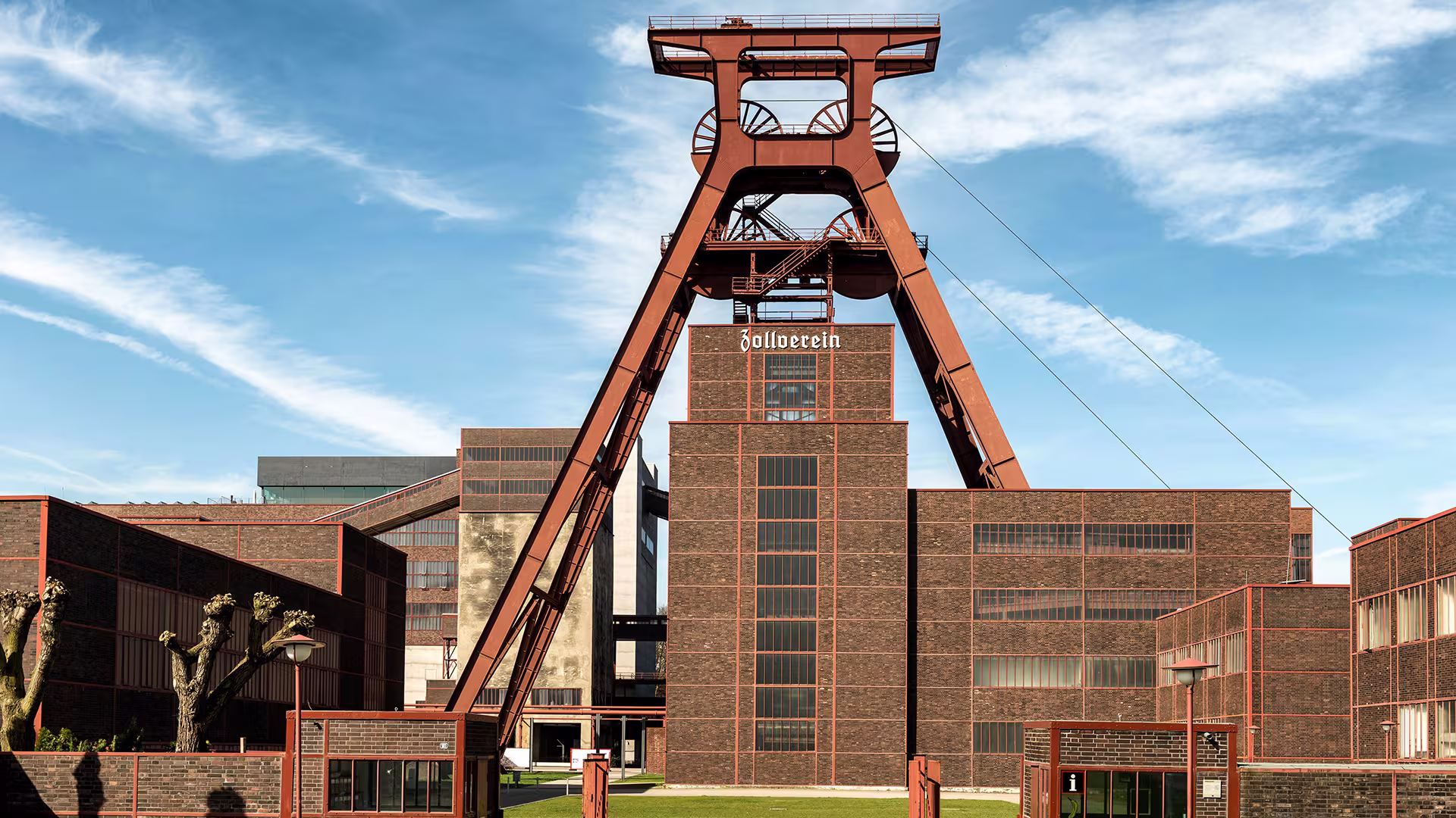 Rotes Doppelbock-Fördergerüst der Zeche Zollverein in Essen über Backsteinbauten, blauer Himmel.