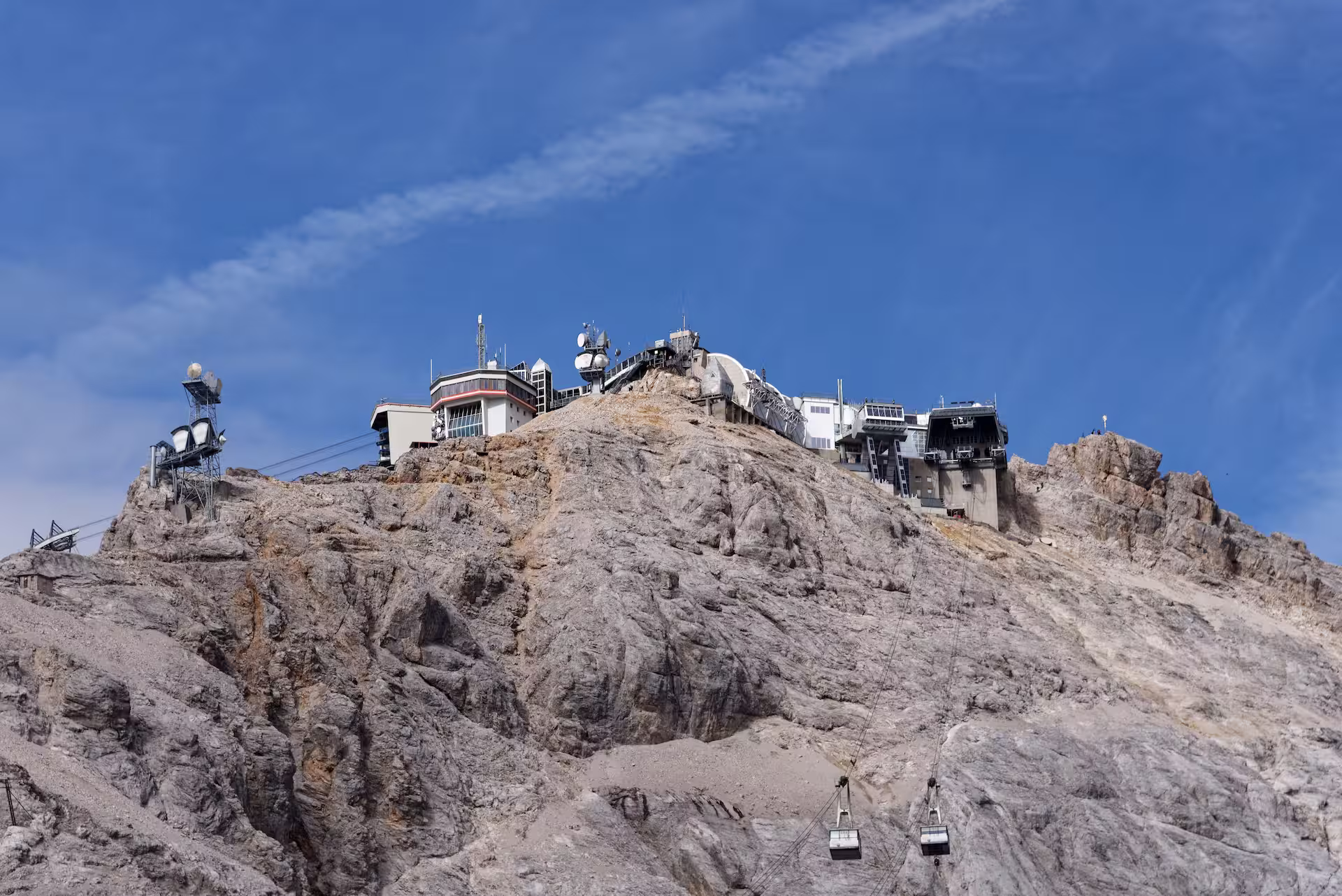 Bebauter Gipfel der Zugspitze mit Seilbahnstationen und zwei Gondeln am Felshang vor blauem Himmel.